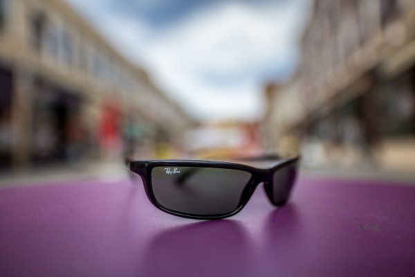 My beat up old Ray-Bans sit on a bistro table, a few inches from the front of the lens, with the buildings flanking either side of a pedestrian mall, rendered soft and abstract at F1.6. 1/4000 F1.6 ISO 200.